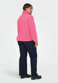 Różowa kurtka polarowa z wysokim kołnierzem, tkaniną o fakturze i opuszczonymi ramionami, zestawiona z ciemnymi jeansami denimowymi i czarnymi butami.