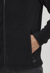 Chaqueta de forro polar negra con una textura suave, cremallera completa, bolsillos laterales y una etiqueta en el dobladillo inferior. Presenta un diseño de muñeca ajustada.