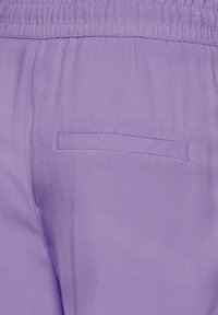 Jasnofioletowe spodnie z elastycznym paskiem w talii i małą tylną kieszenią. Tkanina ma gładką fakturę i delikatny połysk.