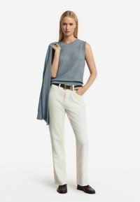 Ärmlös blå stickad topp med texturerat mönster, vita raka byxor, mörkbrunt bälte och svarta loafers. Tröja draperad över axeln.