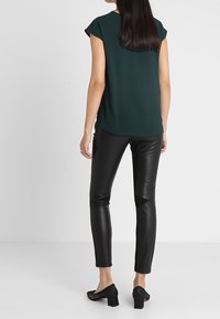 Blusa corta a maniche corte verde scuro con orlo curvo, abbinata a pantaloni in pelle nera dal design elegante e con dettagli tagliati. Scarpe nere con tacco.