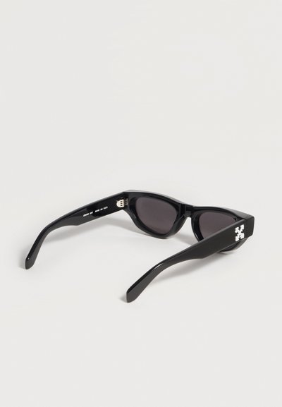 Lunettes de soleil noires œil-de-chat avec verres foncés et logo croix blanche sur la branche, présentées posées sur une surface blanche.