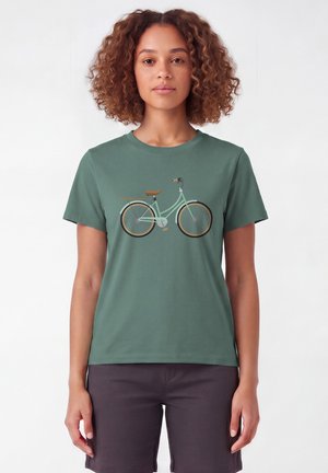 Femme aux cheveux bouclés portant un t-shirt vert avec un motif de bicyclette et un short foncé, debout devant un fond blanc uni.
