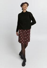 Schwarzer Strickpullover mit hohem Kragen, kombiniert mit einem kastanienbraunen Rock mit weißen Pünktchen. Das Modell trägt schwarze Mokassins und eine Baskenmütze.