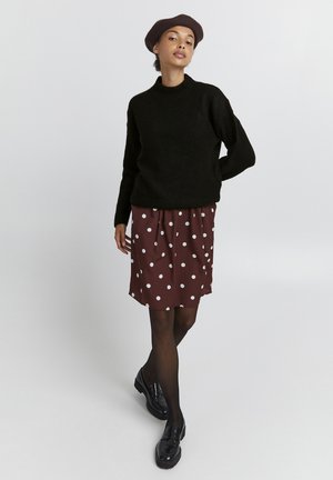 Maglione nero lavorato a maglia con un collo alto, abbinato a una gonna bordeaux con pois bianchi. La modella indossa mocassini neri e un berretto.