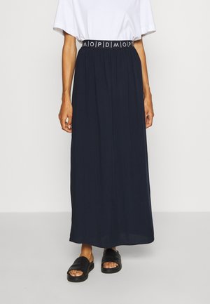 Marineblå maxi nederdel med rynket design og en sort elastisk talje med hvid tekst. Kombineret med enkle sorte sandaler.