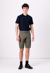 Mornarska polo majica s ovratnikom, u kombinaciji s maslinasto zelenim cargo šortsevima s bočnim džepovima. Model nosi crne čarape i cipele.