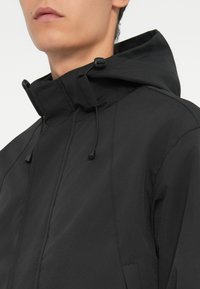 Schwarze Kapuzenjacke mit glatter Textur, ausgestattet mit verstellbaren Kordeln am Hals und einem eleganten Design ohne sichtbare Verschlüsse.