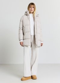 Helle beige Steppjacke mit Kapuze, die ein gestepptes Design, einen vollständigen Reißverschluss und gerippte Bündchen aufweist, kombiniert mit einem weißen gerippten Pullover und hellen Hosen.