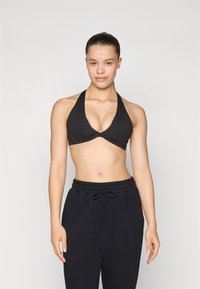 Crni halter bikini top s dizajnom s uvrnutim prednjim dijelom. Mekani materijal, glatka tekstura, minimalna pokrivenost. Uparen s crnim duksericama s vezicom.