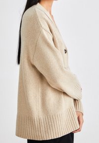 Beige pletený cardigan s výstřihem do V, ribovaným lemem a dlouhými rukávy. Na přední straně jsou dvě černé knoflíky. Jemná textura a volný střih.