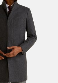 Homme portant un manteau formel gris foncé par-dessus une chemise blanche et une cravate rouge foncé, ajustant le manteau avec ses deux mains.
