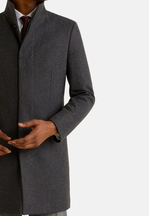 Homme portant un manteau formel gris foncé par-dessus une chemise blanche et une cravate rouge foncé, ajustant le manteau avec ses deux mains.