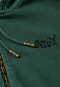 Sudadera verde con cremallera, tela texturizada, logo bordado y detalles metálicos en los cordones con grabado "SUPERDRY".