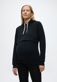 Raskaana oleva nainen, jolla on musta lyhyt huppari valkoisilla kiristysnaruilla ja mustat legginsit, seisoo yksinkertaisen valkoisen taustan edessä.