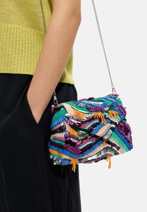 Kolorowa torebka na ramię ozdobiona koralikami i cekinami z srebrnym łańcuszkiem, noszona na ramieniu, zestawiona z żółtą dzianinową bluzką i czarnymi spodniami.