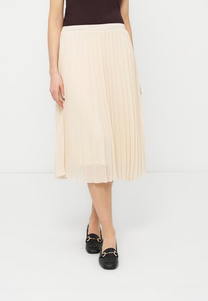 Žena v krémové plisované midi sukni, černém tílkovém topu a černých mokasínách se zlatými přezkami stojí na jednobarevném bílém pozadí.