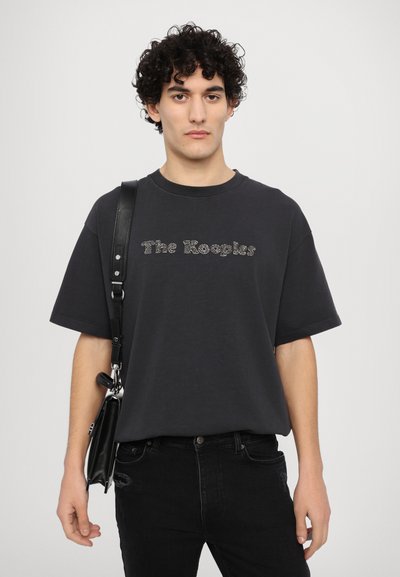 Camiseta oversized negra con el logo de "The Kooples" en un diseño texturizado plateado; combinada con jeans negros y un bolso bandolera negro con herrajes plateados.