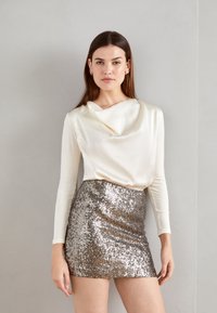 Langes ärmelloses cremefarbenes Oberteil mit drapiertem Ausschnitt, kombiniert mit einem kurzen, metallisch silbernen Paillettenrock, der eine enganliegende Silhouette und eine glatte Textur bietet.