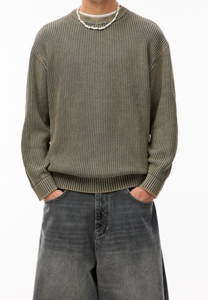 Personne portant un pull côtelé vert olive et un jean noir délavé et ample, les mains dans les poches, accessoirisée d’un collier de perles blanc.