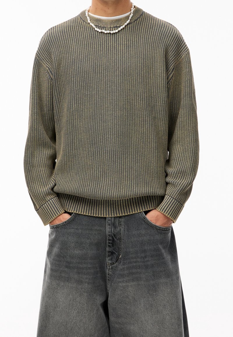 Personne portant un pull côtelé vert olive et un jean noir délavé et ample, les mains dans les poches, accessoirisée d’un collier de perles blanc.