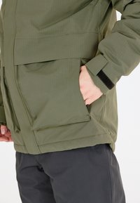 Olivengrüne wasserdichte Jacke aus strukturiertem Stoff, mit großen Fronttaschen, verstellbaren Ärmeln und einem robusten Design mit vertikalen Streifen.