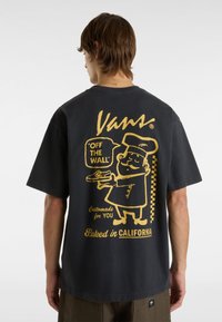 Černé bavlněné tričko s velkým žlutým potiskem na zádech znázorňujícím animovaného pekaře a textem "Vans" a "Baked in CALIFORNIA."
