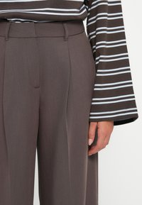 Brown tailored trousers with a smooth texture and front pleats, worn with a long-sleeve, striped shirt featuring blue and white lines.