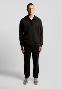 Zwarte zip-up hoodie met een getextureerde capuchon en een borstpaneel, gecombineerd met zwarte broek en lichtgrijze sneakers. Eenvoudig, atletisch ontwerp.