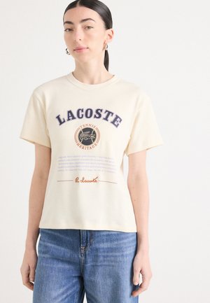 T-shirt crema con grafica "LACOSTE" in blu navy, con "TENNIS HERITAGE" sottostante. Maniche corte, vestibilità rilassata, texture morbida. Jeans in denim visibili.