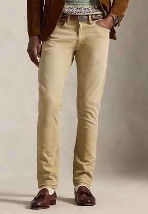 Man met beige slim-fit corduroy broek, bruine loafers met kwastjes, gebreide trui met patroon en bruine suède blazer, met één hand in de zak.