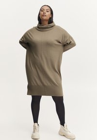 Robe en tricot olive surdimensionnée avec un col drapé, des manches tombantes et une coupe droite; associée à des collants noirs et des baskets beiges.