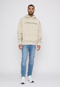 Beige hætte sweatshirt med frontlomme, der har sort "PEGAHOR" tekst. Bæres med lyseblå slim-fit jeans og hvide sneakers.