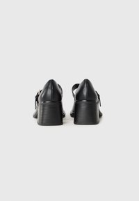 Pair of black leather block-heeled shoes with ankle straps and silver buckles, viewed from the back on a plain white background.