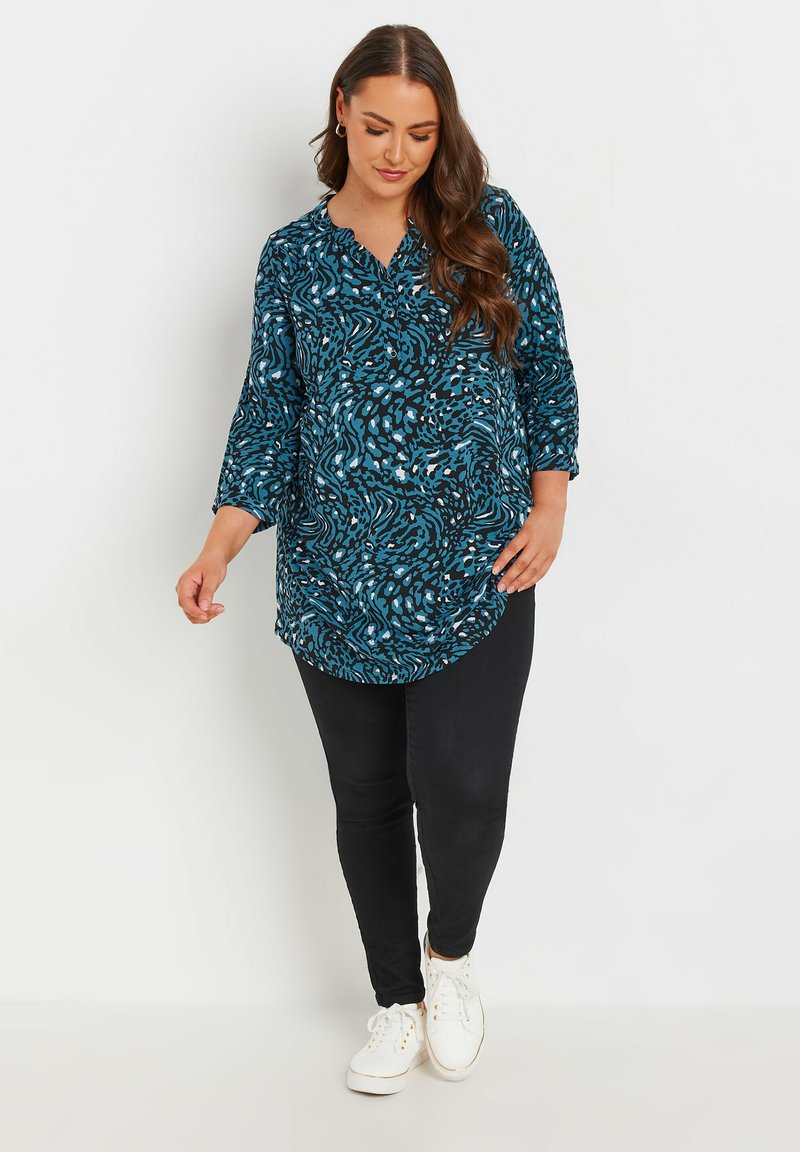 Camicia lunga con un motivo astratto verde e nero, davanti con bottoni e maniche a tre quarti, abbinata a jeans skinny neri e scarpe da ginnastica bianche.
