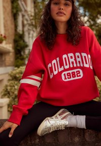 Červená mikina s nápisem "COLORADO 1982", bílé pruhy na rukávech, kombinovaná s černými legínami a lesklými stříbrnými teniskami.