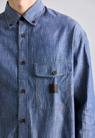 Bărbat îmbrăcat cu o cămașă din denim albastru, cu nasturi, având un buzunar pe piept și cusături maro vizibile, pe un fundal deschis.