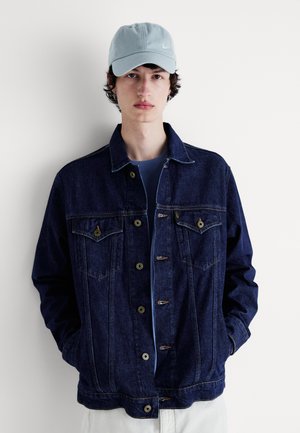 Dunkelblaue Jeansjacke mit Knopfverschluss, zwei Brusttaschen und kontrastierenden Nähten. Wird über einem blauen Hemd getragen und mit einer hellblauen Mütze kombiniert.
