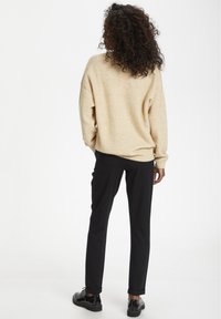 Pull en tricot beige et pantalon noir ajusté. Le pull présente une coupe décontractée et des poignets côtelés. Chaussures noires avec une finition lisse.