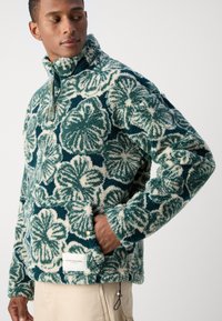 Pullover in pile con motivo floreale teal e crema, colletto alto, zip frontale e tasche laterali; tessuto morbido e testurizzato.