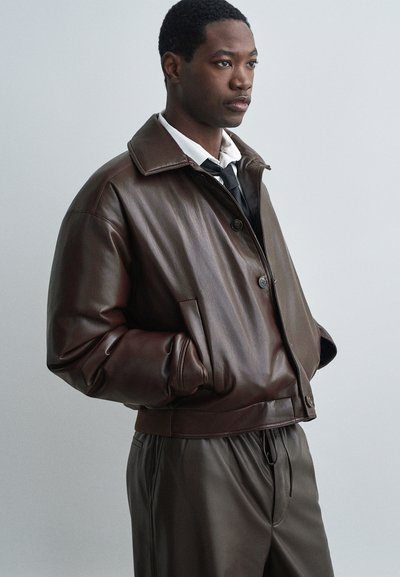 Jachetă bomber din piele maro, cu un guler lat, închidere cu nasturi și buzunare laterale. Purtați cu pantaloni maro asortați și o cămașă albă dedesubt.