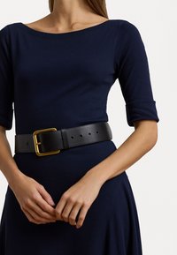 Black leather belt with a large gold buckle, worn over a dark blue garment with a wide neckline and three-quarter sleeves.