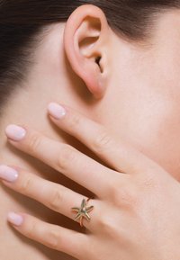 Anillo de estrella de mar dorada con un detallado texturizado, llevado en un dedo delgado. El fondo presenta piel y una oreja con tonos neutros.