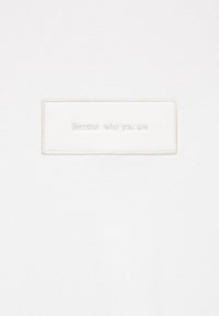 Tissu blanc avec un patch rectangulaire cousu comportant le texte brodé en fil clair « Deviens qui tu es ».