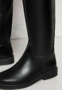 Botas de cuero negras hasta la rodilla con una textura suave, puntera redonda, costuras mínimas y un tacón bajo en bloque. Presentan un diseño elegante y ajustado.