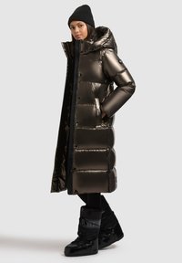 Lang, puffy jas in metallic donkerbruin met een capuchon, voorzien van grote geweven secties en zijzakken. Zwarte, geïsoleerde laarzen maken de look compleet.