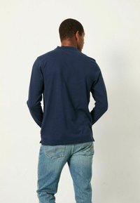 Marineblaues Langarmhemd aus strukturiertem Stoff, mit abgerundetem Saum und seitlichen Schlitzen. Getragen mit hellblauen Jeans, die eine lässige Passform zeigen.