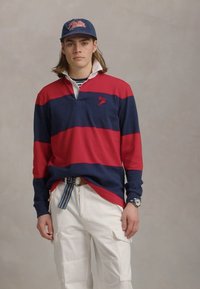 Polo à rayures rouge et marine à manches longues avec un col blanc, associé à un pantalon cargo blanc et une casquette bleue. Il est doté d'une ceinture multicolore.