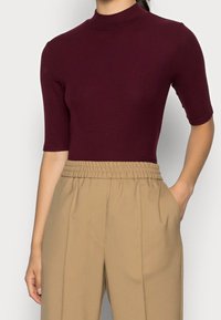 Žena v těsném vínovém topu s polovičními rukávy, zasunutém do vysokých béžových kalhot s elastickým pasem a postranními kapsami.