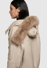 Beige vadderad parka med avtagbar huva i mjuk brun pälsimitation och sidofickor med dragkedja. Slät textur med en något glänsande finish.